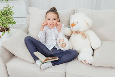 Kanepede yanında oyuncak ayıyla oturan, peri masalı okuyan, dişlerini döken, canavar taklidi yapan tatlı küçük bir kız..