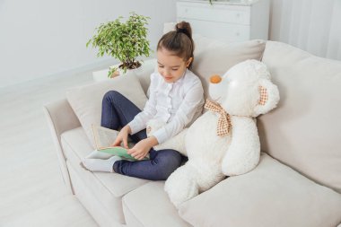 Kanepede oturan sevimli küçük kız yanında oyuncak ayıyla peri masalı okuyor..