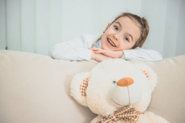 Büyük beyaz ayı kanepede. Kız kanepenin arkasında duruyor, sırtına yaslanıyor, ellerini kavuşturuyor, kameraya bakıyor, hoş bir şekilde gülümsüyor..