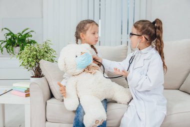 Oyunu oynayan meraklı kızlar: doktor ve hasta. Küçük doktor steteskopla hastasının kalp atışlarını dinliyor. Tıbbi maskeli Teddy kızların dizlerinin üstünde..