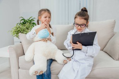 Beyaz ayıcık hasta. Oldukça küçük bir doktor, peluş hastası için ilaç yazıyor. Başka bir kız korkmuş bir ifadeyle oturuyor..