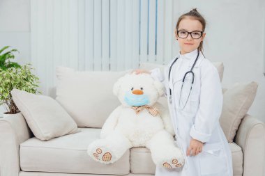 Doktor önlüğü, steteskop ve gözlük takan, tıbbi maskeli oyuncak ayının kafasında duran, el ele tutuşan kendine güvenen küçük bir kız..