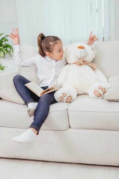 Linda niña sentada en el sofá con osito de peluche cerca de ella, leyendo cuento de hadas . — Foto de Stock