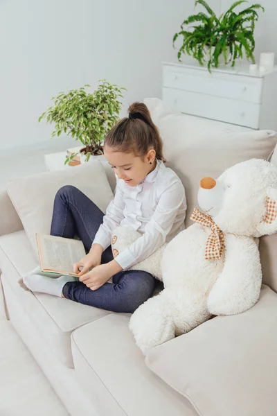 Симпатична дівчинка сидить на дивані з плюшевим ведмедем біля неї, читаючи казку . — стокове фото