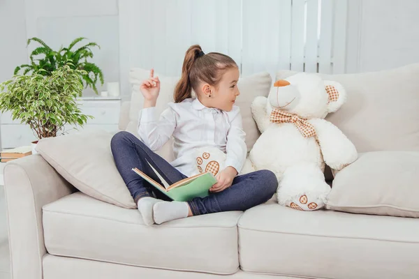 Niza niña sentada en el sofá con osito de peluche cerca de ella, leyendo cuento de hadas, señalando con el dedo hacia arriba, diciendo algo importante a su amigo suave . — Foto de Stock