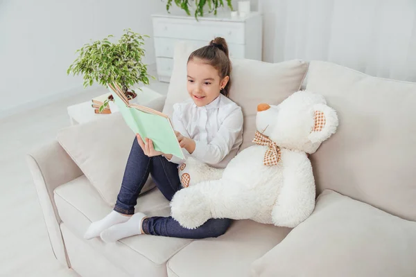 Cute little girl sitting on the sofa with teddy-bear near her, reading fairy-tale, pointing her finger at something in the book, smiling.