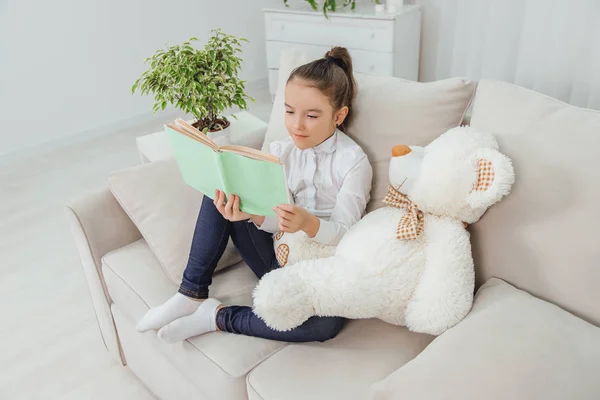 Cute little girl sitting on the sofa with teddy-bear near her, reading fairy-tale.