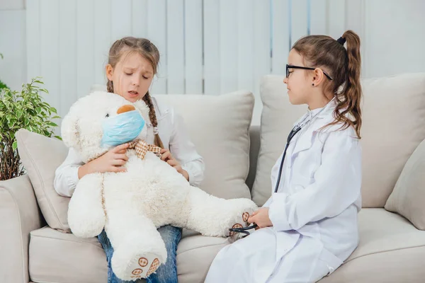 Two funny girls playing the game of doctor and patient. One girl is holding an ill teddy-bear in medical mask on her knees. Another girl is s doctor. — Stock Photo, Image