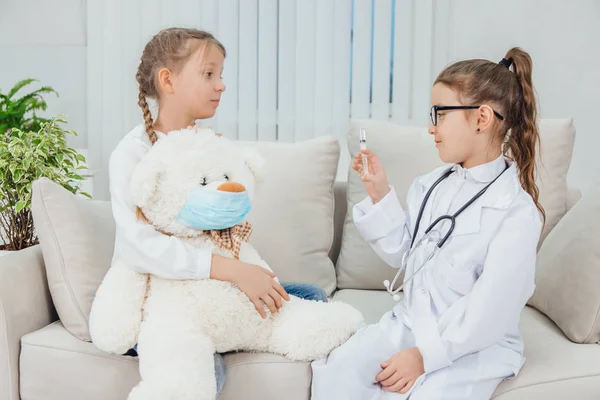 Schattig klein meisje in doktersuniform houdt een spuit vast, maakt van witte teddybeer een schot. Een ander meisje zit, houdt handen op de wangen, kijkt bezorgd en bang. — Stockfoto