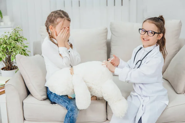 Schattig klein meisje in doktersuniform maakt van witte teddybeer een shot. Een ander meisje zit, kijkt bezorgd en bang, bedekt haar gezicht met handen. — Stockfoto