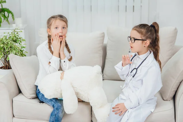 Cute little girl in doctor uniform is holding a syringe, going to make white teddy-bear a shot. Another girl is sitting, keeping hands on the cheeks, looking worried and scared. — Stock Photo, Image