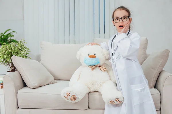 Niña bastante sorprendida vistiendo abrigo médico, estetoscopio y gafas, de pie, cogida de la mano en la cabeza de osito de peluche en máscara médica . —  Fotos de Stock