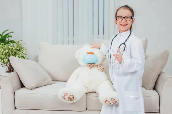 Niña bastante sonriente que lleva abrigo médico, estetoscopio y gafas, de pie, cogida de la mano en la cabeza del osito de peluche con máscara médica. El chico señala con el dedo hacia arriba . —  Fotos de Stock