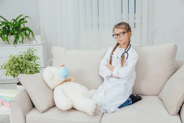 Bonita niña con abrigo médico, estetoscopio y gafas, sentada en el sofá con las piernas acurrucadas, las manos dobladas, mirando a la cámara. El oso de peluche enfermo está tendido cerca de ella. . —  Fotos de Stock