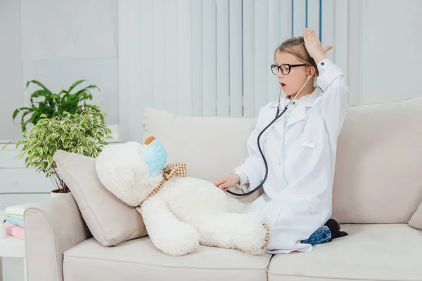 Niña bonita vistiendo abrigo médico, sentada en el sofá con las piernas acurrucadas, examinando a su oso de peluche enfermo con estetoscopio . —  Fotos de Stock