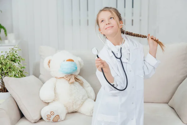 Bastante confiada niña usando abrigo médico, sosteniendo estetoscopio, tirando de su hermosa trenza, de pie, posando . —  Fotos de Stock