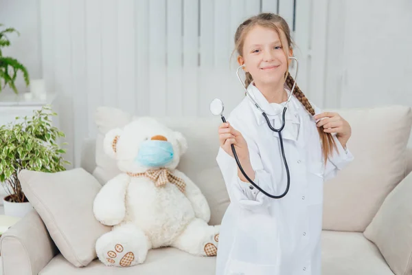 Bastante confiada niña usando abrigo médico, sosteniendo estetoscopio, tirando de su hermosa trenza, de pie, posando . —  Fotos de Stock