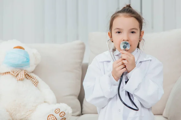 La niña con abrigo médico está hablando en el estetoscopio. Osito de peluche blanco con máscara médica está sentado cerca de ella . —  Fotos de Stock