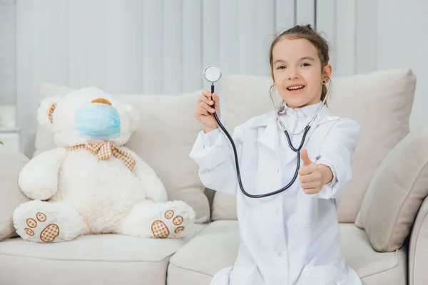 Niña con abrigo médico sosteniendo un estetoscopio, apuntando con el dedo hacia arriba. Osito de peluche blanco con máscara médica está sentado cerca de ella . —  Fotos de Stock