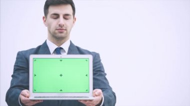 Gri takım elbiseli ve kravatlı yakışıklı bir iş adamı, ayakta duruyor, elinde yeşil ekranlı bir laptop tutuyor, başını sallıyor, onaylıyor..