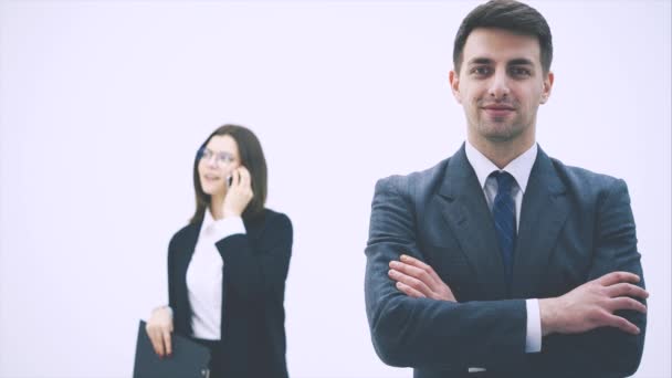 Guapo hombre de negocios sonriente de pie en la vanguardia, con las manos cruzadas. Mujer en el fondo está hablando por teléfono . — Vídeos de Stock