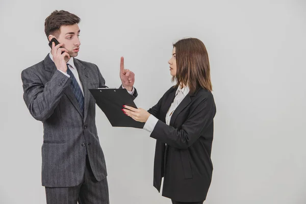 Handsome busy businessman is talking on the phone, pointing his finger up for his secretary to wait. Secretery standing with clipboard, waiting for bosss signature. — Stock Photo, Image