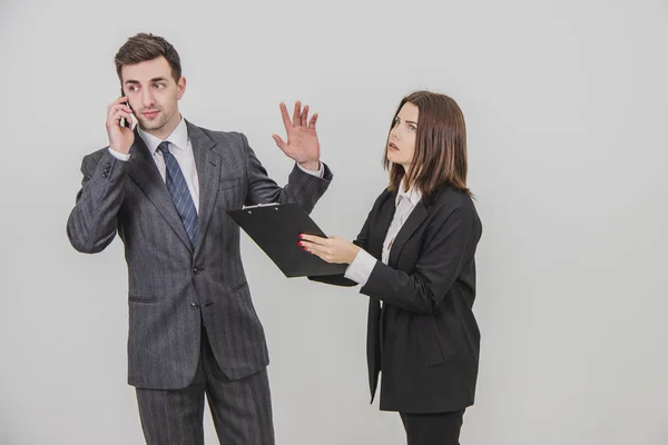 Un hombre de negocios guapo y ocupado está hablando por teléfono, señalando con el dedo a su secretaria para que espere. Secreto de pie con portapapeles, esperando la firma del jefe . — Foto de Stock