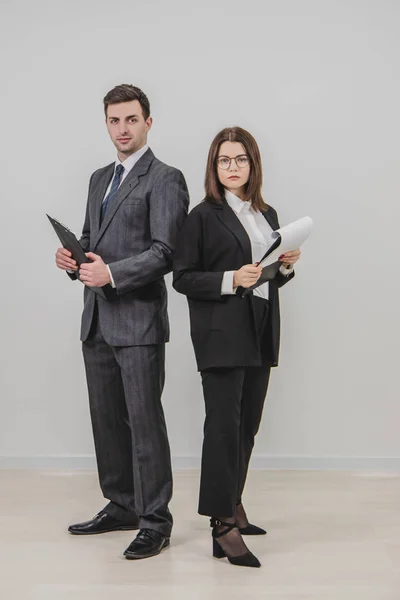 Full-length business man and woman standing shoulder to shoulder, holding clipboards, looking confidently at the camera. — Stock Photo, Image