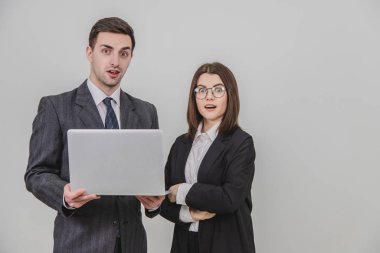 İş ortakları ayakta duruyor, laptoptaki verileri kontrol ediyor, hayret verici yüz ifadeleriyle kameraya bakıyor. Kadın ayakta, elleri bağlı..