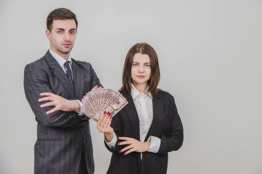Zengin, kendine güvenen bir iş çifti. Adam elleri bağlı ayakta duruyor. Onun tarafındaki kadın bir dolar banknotunu tutuyor..