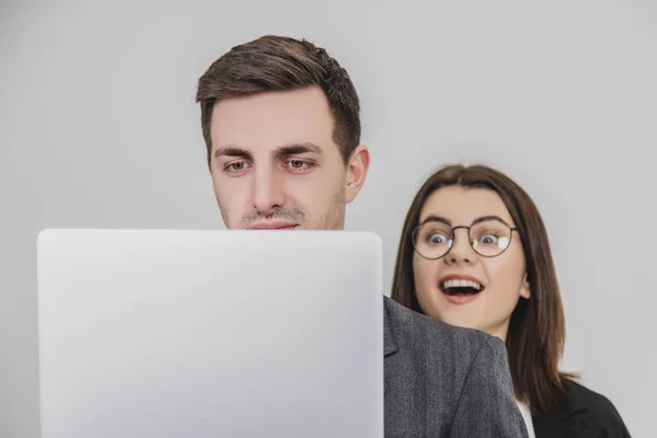 Biznesmen stoi, pracuje z laptopem. Dociekliwa kobieta patrzy mu przez ramię na ekran, jej ciekawy oczy szeroko otwarte. — Zdjęcie stockowe