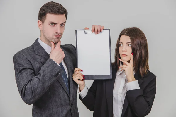 Successful business couple standing with thoughtful face expressions, holding together a clipboard with empty clear sheet of paper for text, thinking. — Stock Photo, Image