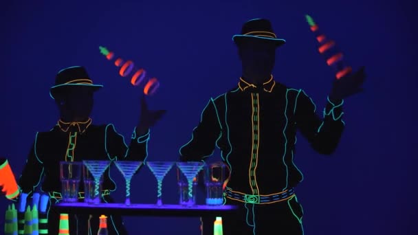 Barmanská show. Výkon. Barmanka a barman spolu žonglují v neonové uniformě na jasně modrém pozadí. Zpomal. 4k. — Stock video