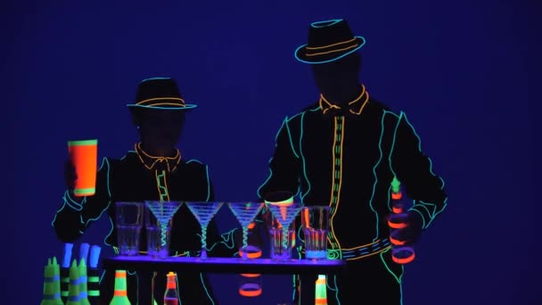 Arrivano i barman. Prestazioni. Barista e barista stanno versando alcol insieme, vestiti in uniforme al neon su sfondo blu brillante. Al rallentatore. 4K . — Video Stock