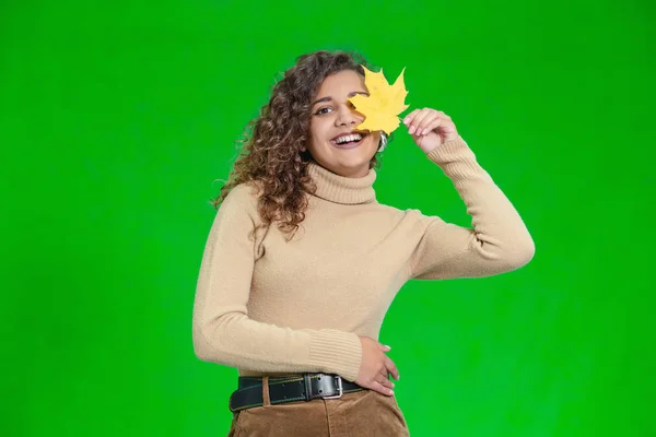 Χαμογελαστή γυναίκα κρατώντας στα χέρια της κίτρινο φύλλο σφενδάμου μπροστά από το μάτι της, στέκεται πάνω σε πράσινο φόντο. — Φωτογραφία Αρχείου