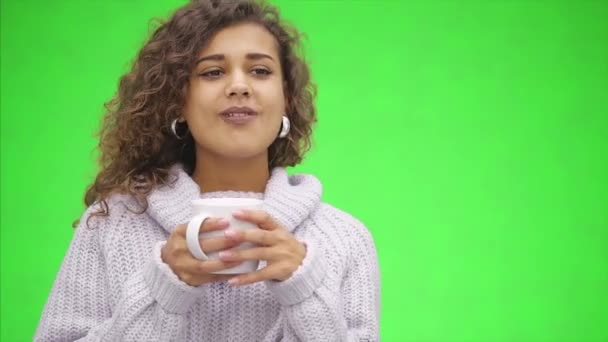 Una chica afroamericana atractiva está masticando. Ella se está calentando sosteniendo una taza de bebida caliente, usando suéter blanco. De cerca. Copiar espacio . — Vídeos de Stock