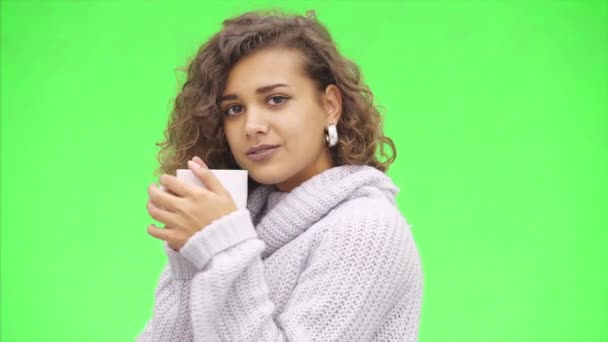 Attraente ragazza afro-americana sta tenendo una tazza bianca ampiamente. Si sta scaldando, indossa un maglione bianco. Chiudete. Copia spazio . — Video Stock