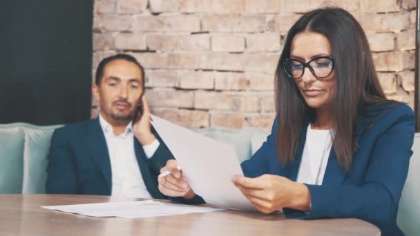 Dos empresarios trabajan en un café. La mujer está aprendiendo atentamente los documentos. El hombre está al teléfono, en un fondo borroso. De cerca. Copiar espacio . — Vídeos de Stock