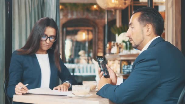 Zwei Geschäftsleute arbeiten in einem Café. Frau lernt aufmerksam die Dokumente. Mann ist im Videochat. Seitenansicht. Nahaufnahme. Kopierraum. — Stockvideo