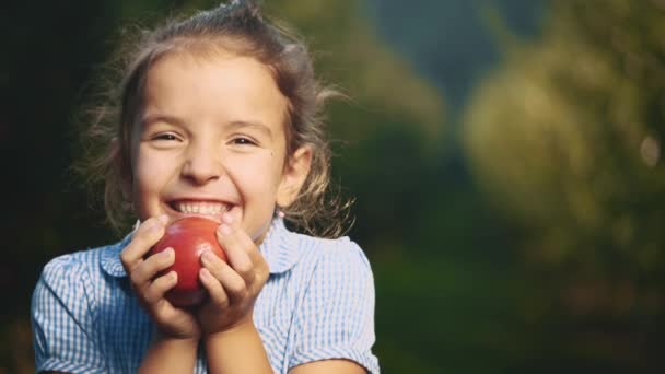 Küçük kız yüzüne yakın kırmızı bir elma tutuyor. Kız herkese gülümsüyor. Kapatın. Uzayı kopyala 4k. — Stok video