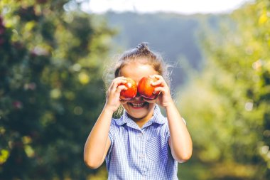 Mavi elbiseli, sevimli, gülümseyen bir çocuk, bulanık arka planda elmalarını gözünün önünde tutuyor..