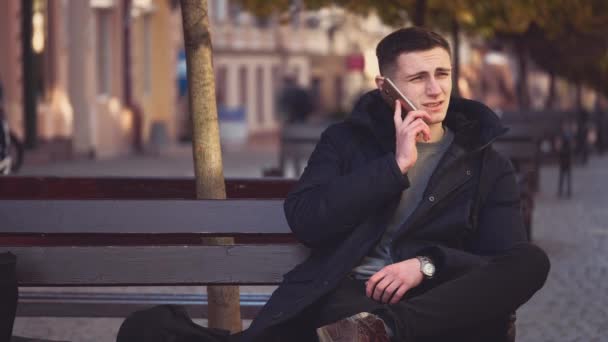 Gutaussehender, attraktiver junger Mann telefoniert und sitzt auf der Bank. Er schaut sich um. Kopierraum. Aktion. Animation. 4k. — Stockvideo