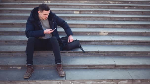 Um jovem está sentado nas escadas e usando seu smartphone. Espaço para cópia. Acção. Animação. 4K . — Vídeo de Stock