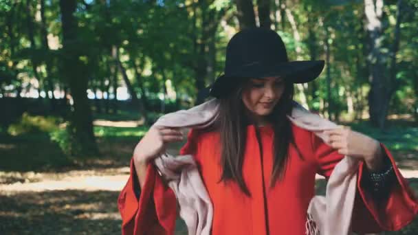 Bonita joven con abrigo rojo y sombrero negro es vinculante bufanda suavemente rosa, aislado, sobre fondo borroso parque. De cerca. Copiar espacio. 4K . — Vídeos de Stock