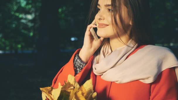 Lovely girl is on the phone, holding a bunch of autumn leaves. Blurred background. Crop. Close up. Copy space. 4K. — Stock Video