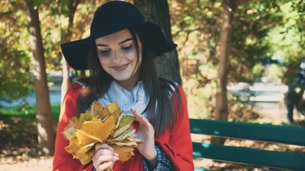 Young lovely girl is holding a bunch of autumn leaves, while sitting on the bench. Blurred background. Close up. Copy space. 4K. — 비디오