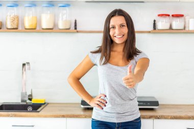 Pretty smiling woman with dark hair looking at the camera, giving thumb up, spending time in her new beautiful cozy kitchen. clipart