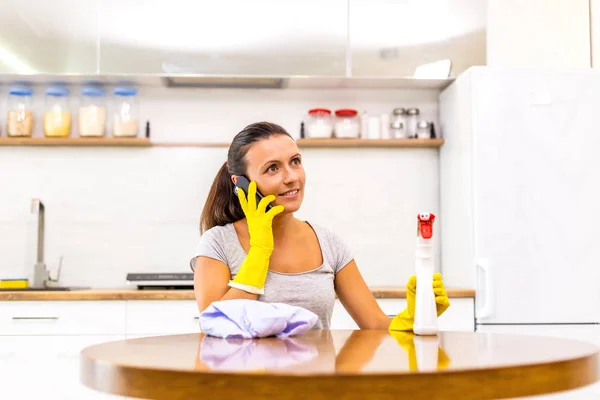 Foto di bella donna vestita in abbigliamento casual che parla sul telefono cellulare mentre lucida il tavolo . — Foto Stock