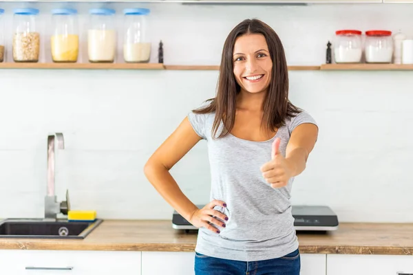 Hübsche lächelnde Frau mit dunklen Haaren, die in die Kamera schaut, den Daumen nach oben gibt, Zeit in ihrer neuen schönen, gemütlichen Küche verbringt. — Stockfoto