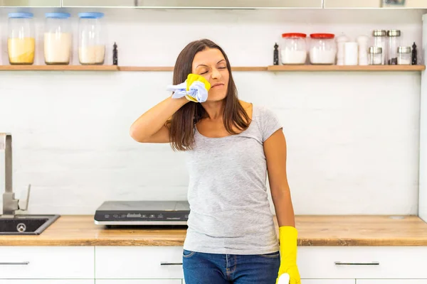 Женщина устала после тяжелой работы по уборке, вытирая пот с лица и держа бутылку спрея на кухонном фоне . — стоковое фото